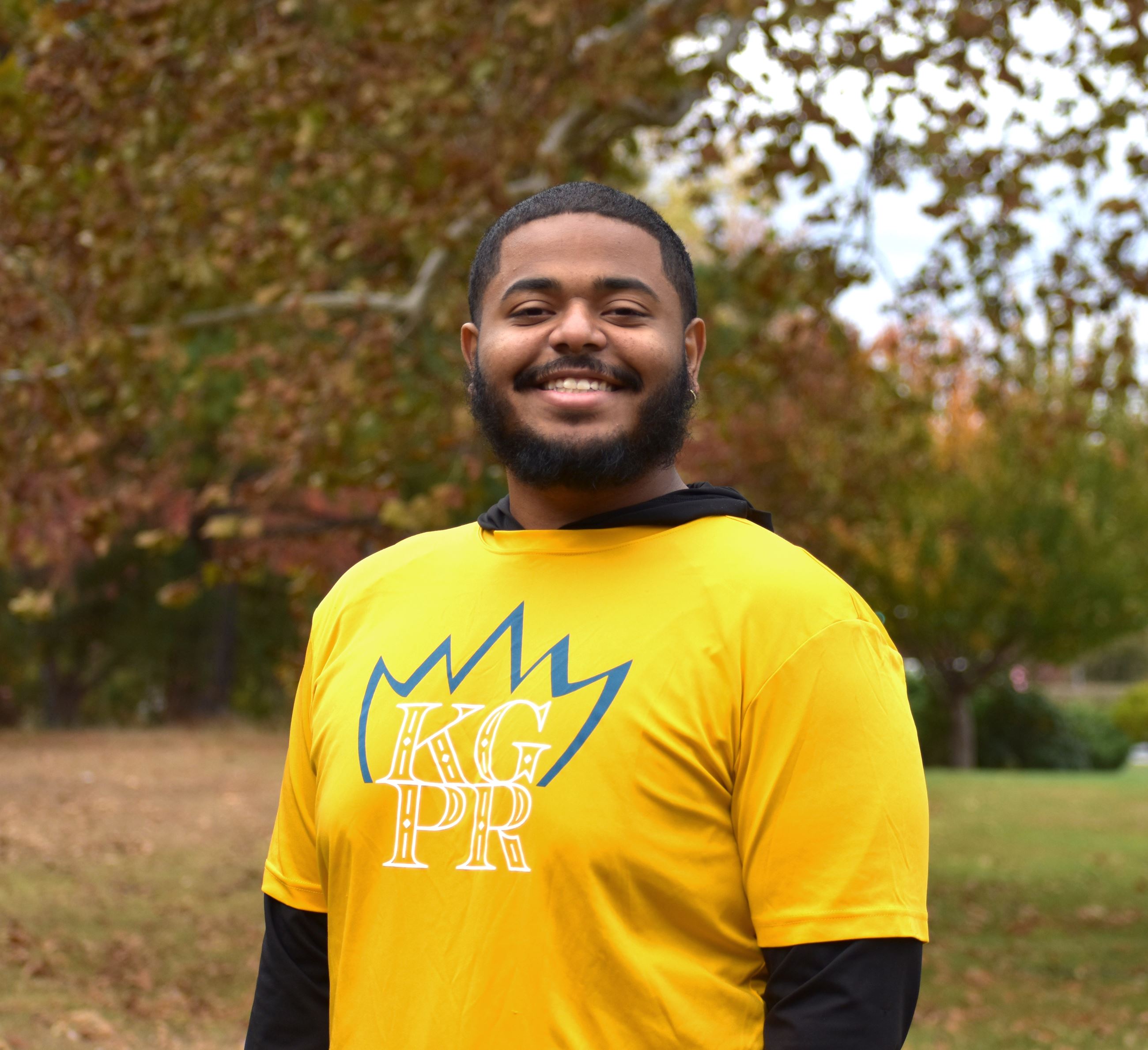 A headshot of a man in a yellow KGPR shirt against a fall background.